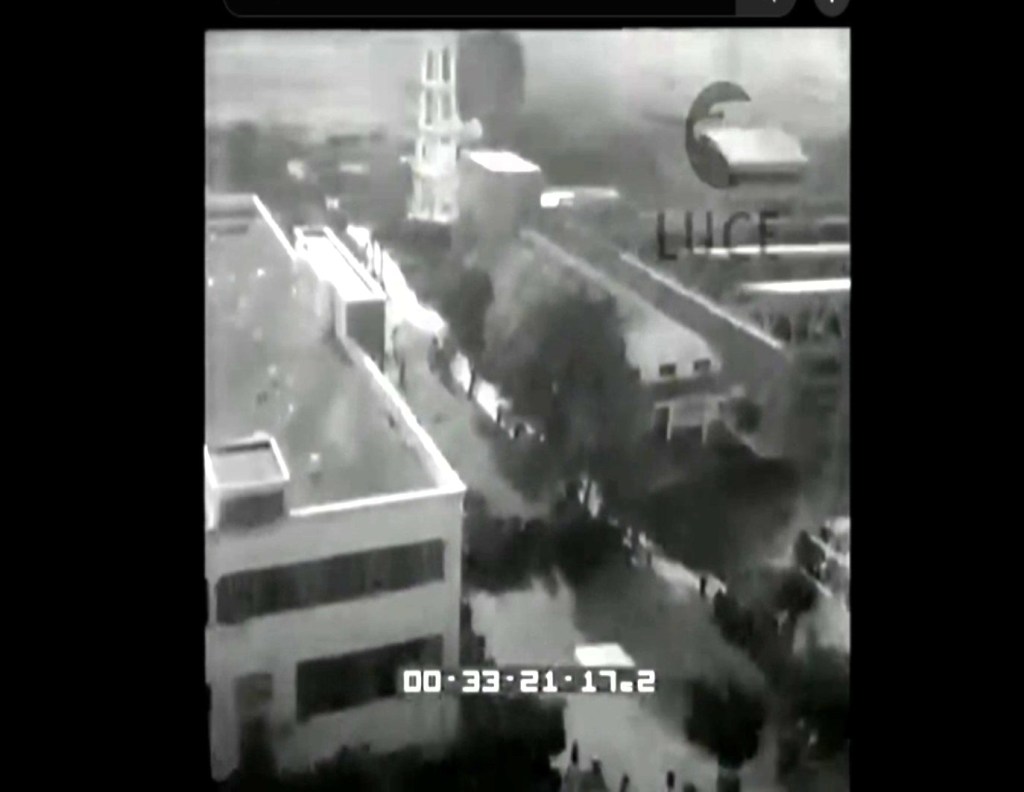 Ripresa in bianco e nero di una strada con un gruppo di persone in movimento, alberi e edifici visibili sullo sfondo.