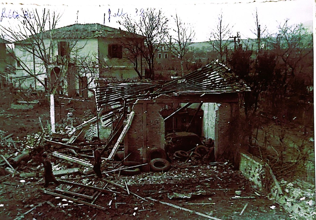 Immagine di un edificio distrutto con un tetto crollato, circondato da detriti e alberi spogli, in un paesaggio triste e desolato.