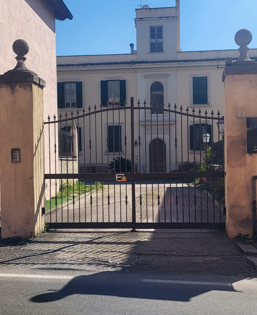 Ingresso con cancello in ferro di una villa, visibile il giardino e l'edificio sullo sfondo sotto un cielo sereno.